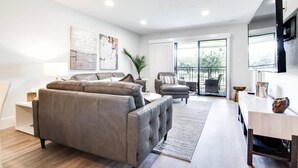 Un salon moderne avec un canapé gris, une petite table et un téléviseur. Il y a un balcon avec vue sur les arbres.