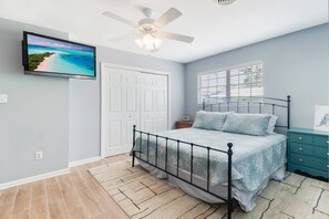 Une chambre à coucher comprenant un lit, un ventilateur de plafond, un téléviseur fixé au mur et une fenêtre avec des stores.