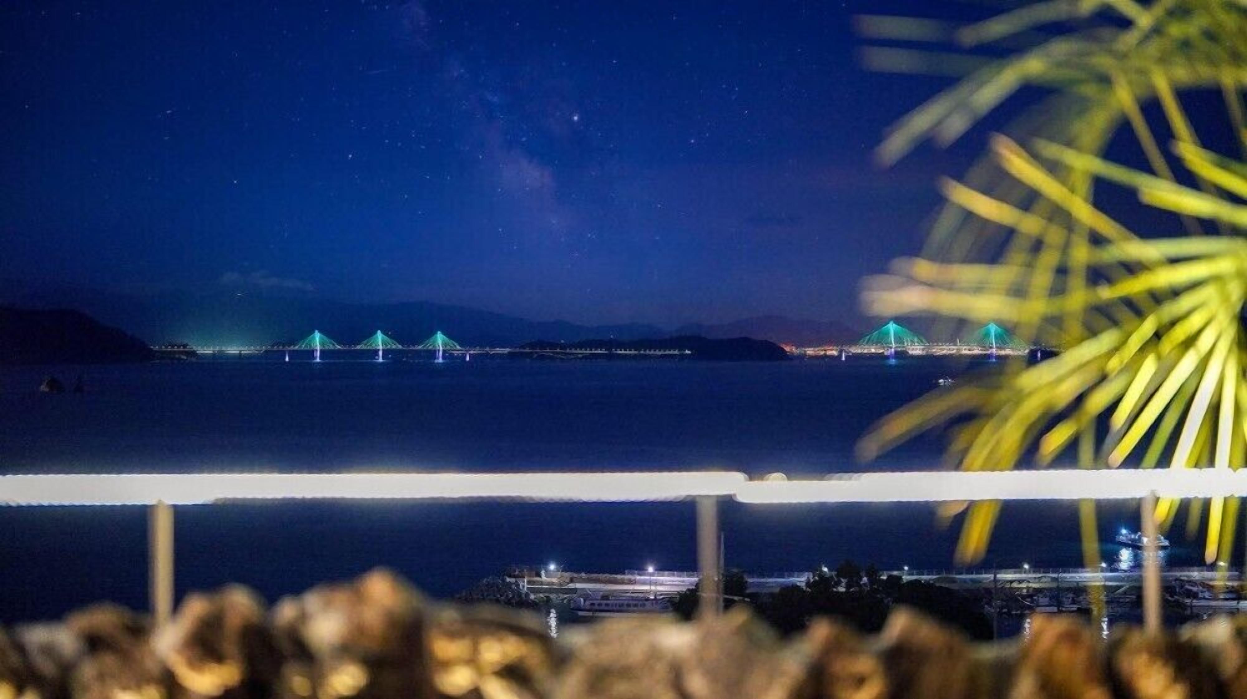 Vista nocturna de un puente con estructuras iluminadas, una masa de agua y una costa distante.