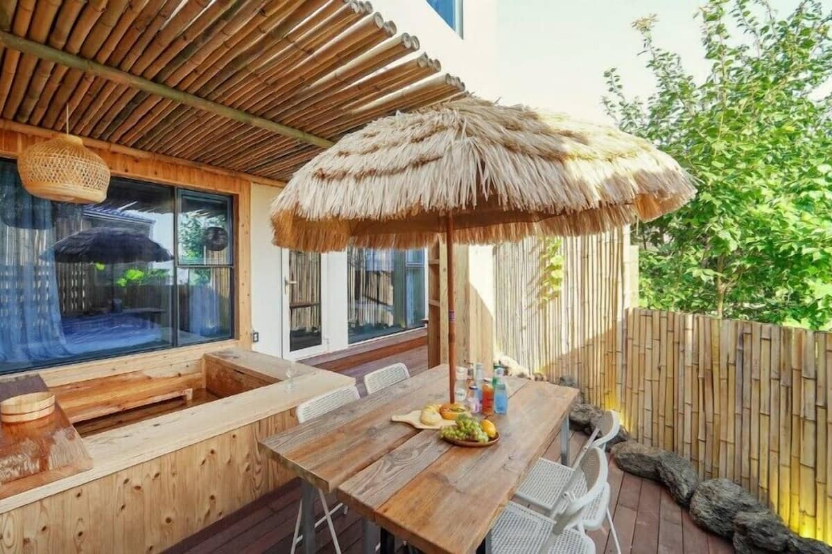 A wooden deck with a table, chairs, a large woven umbrella, and a wooden bench.