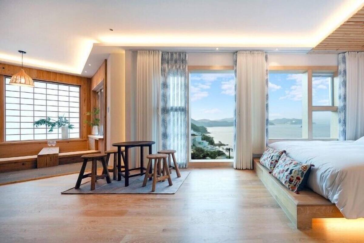 A modern hotel room with a large bed, a dining area with stools, and a view of the sea.