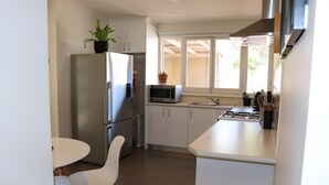 A compact kitchen with white cabinets, a stainless steel refrigerator, a microwave, and a gas stove. There is a small dining table with white chairs. - The Beach House At Henley (Henley Beach)