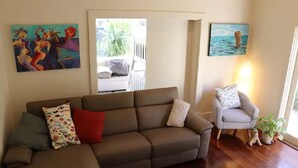 A living room with a brown sectional sofa, a grey armchair, and a small table with a lamp. There are two paintings on the walls and a window with a view of the outdoors. - The Beach House At Henley (Henley Beach)