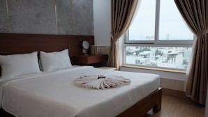 Una cama bien hecha con edredón blanco y un arreglo floral, una mesita de noche de madera con espejo y libros, y un ventanal con cortinas.
