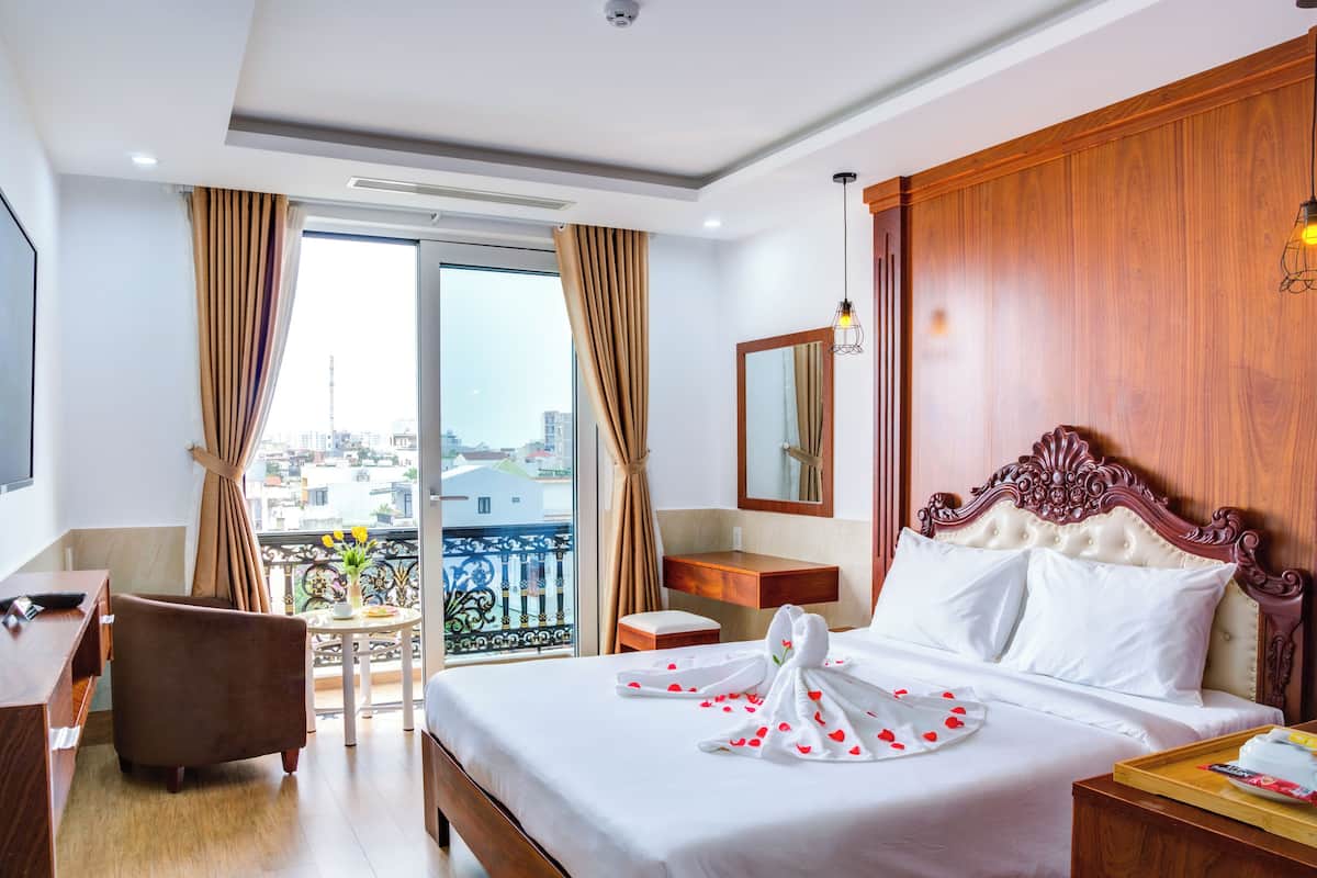 A hotel room with a large bed, a wooden headboard, a desk, a chair, a small table with a vase, and a balcony with curtains.