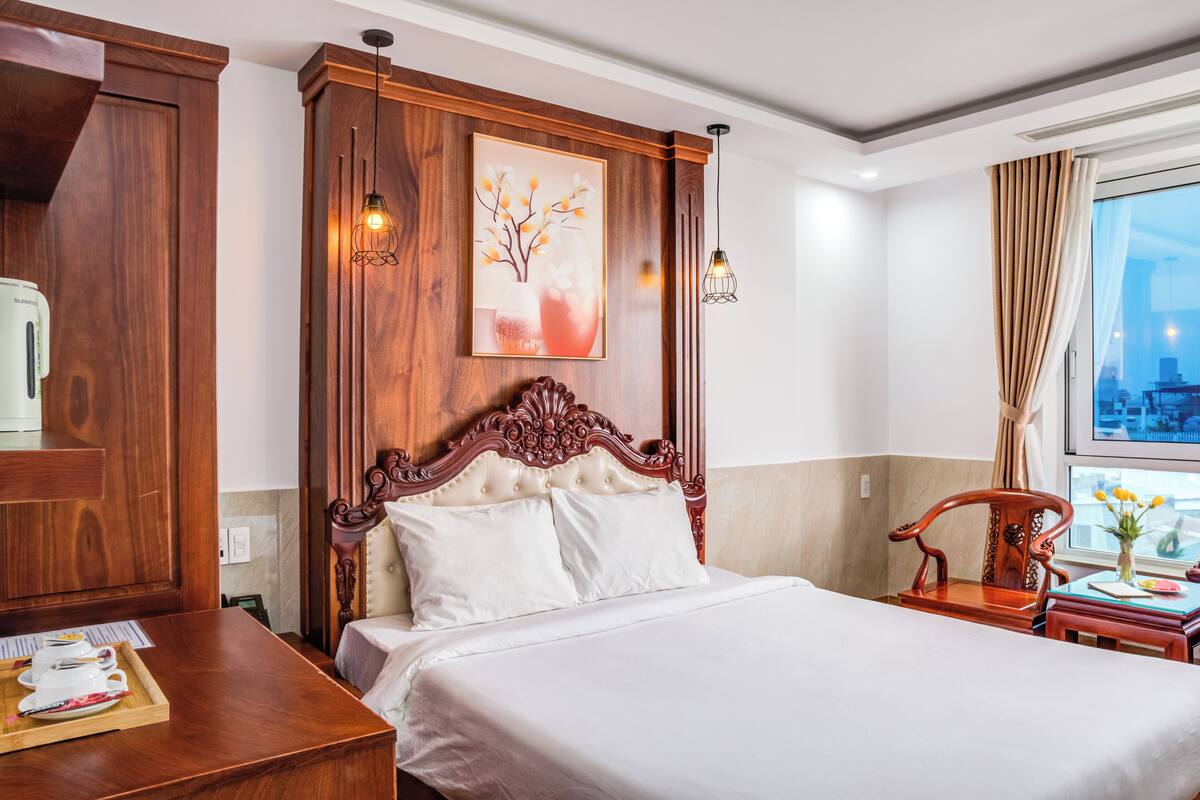 A hotel room with a large bed, wooden headboard, a painting on the wall, a desk with a tray of tea, and a view of the city through the window.