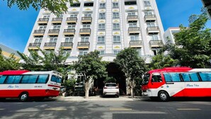 Un edificio de varios pisos con una entrada central, rodeado de árboles y dos autobuses estacionados frente a él.