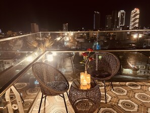 Uma varanda com cadeiras de vime e uma mesa, um vaso com flores, e vista para a cidade à noite.