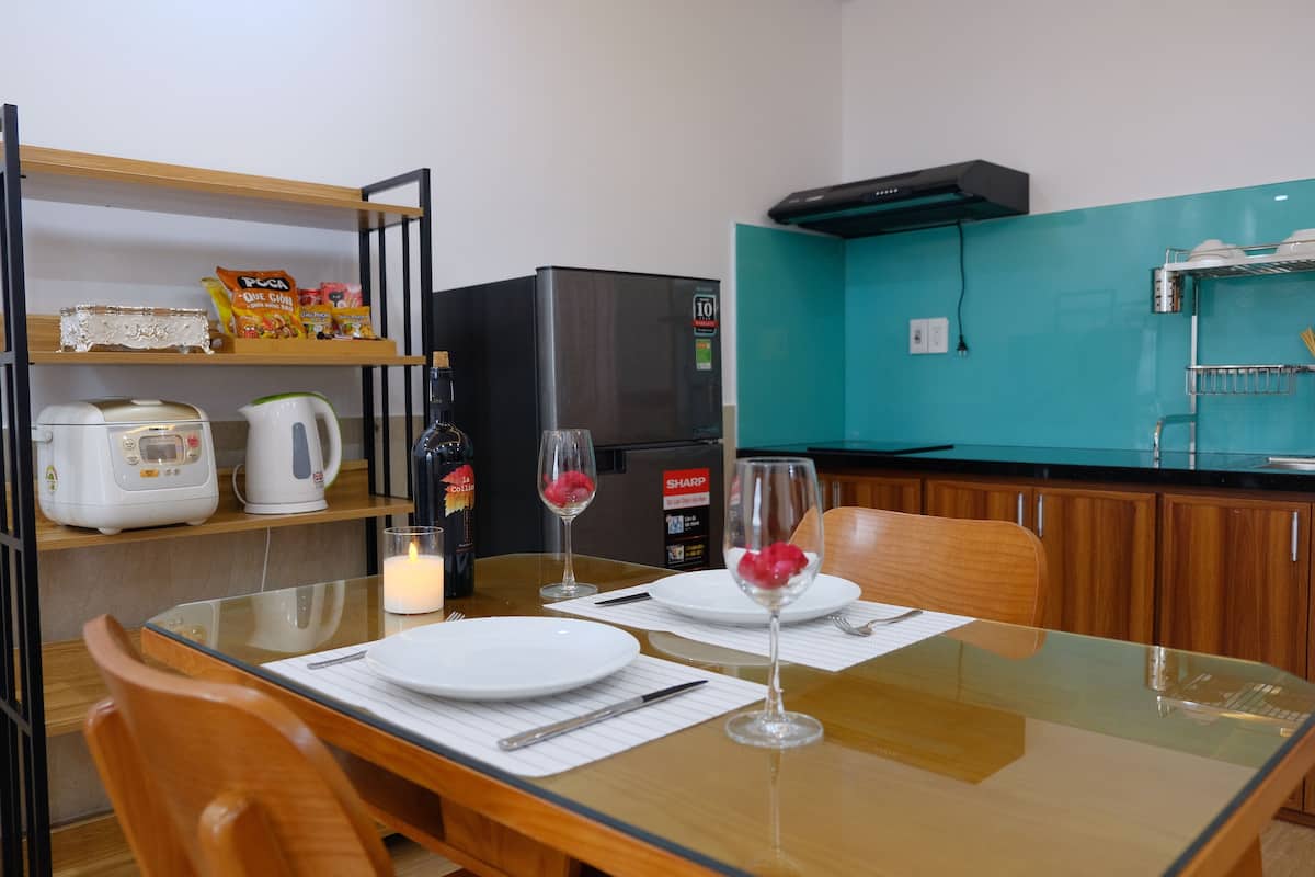 A dining area with a glass table set for two, a refrigerator, a coffee maker, and a shelf with snacks.