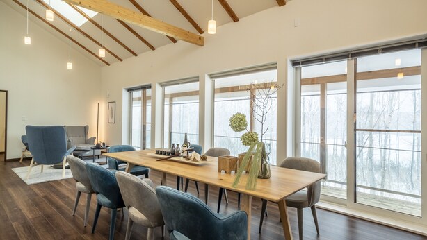 A spacious room with a long wooden table, blue chairs, and large windows offering a view of trees.