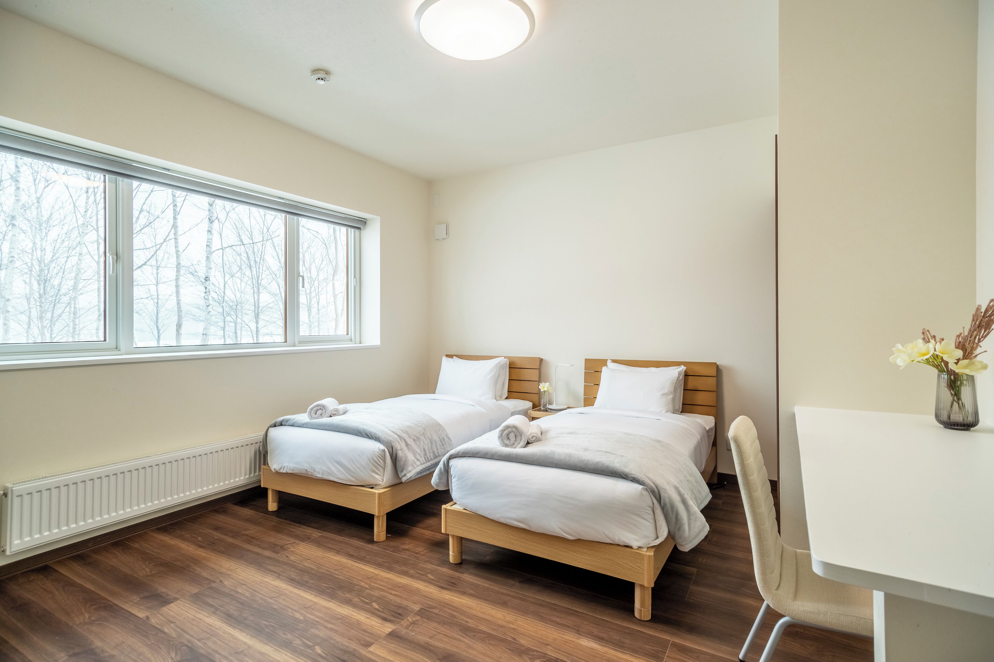 A room with two single beds, a white desk, and a window with a view of bare trees.
