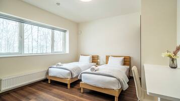 A room with two single beds, a white desk, and a window with a view of bare trees.