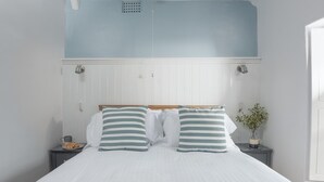 Een slaapkamer met een bed, twee kussens, een nachtkastje, een kleine tafel, een plant en een raam.