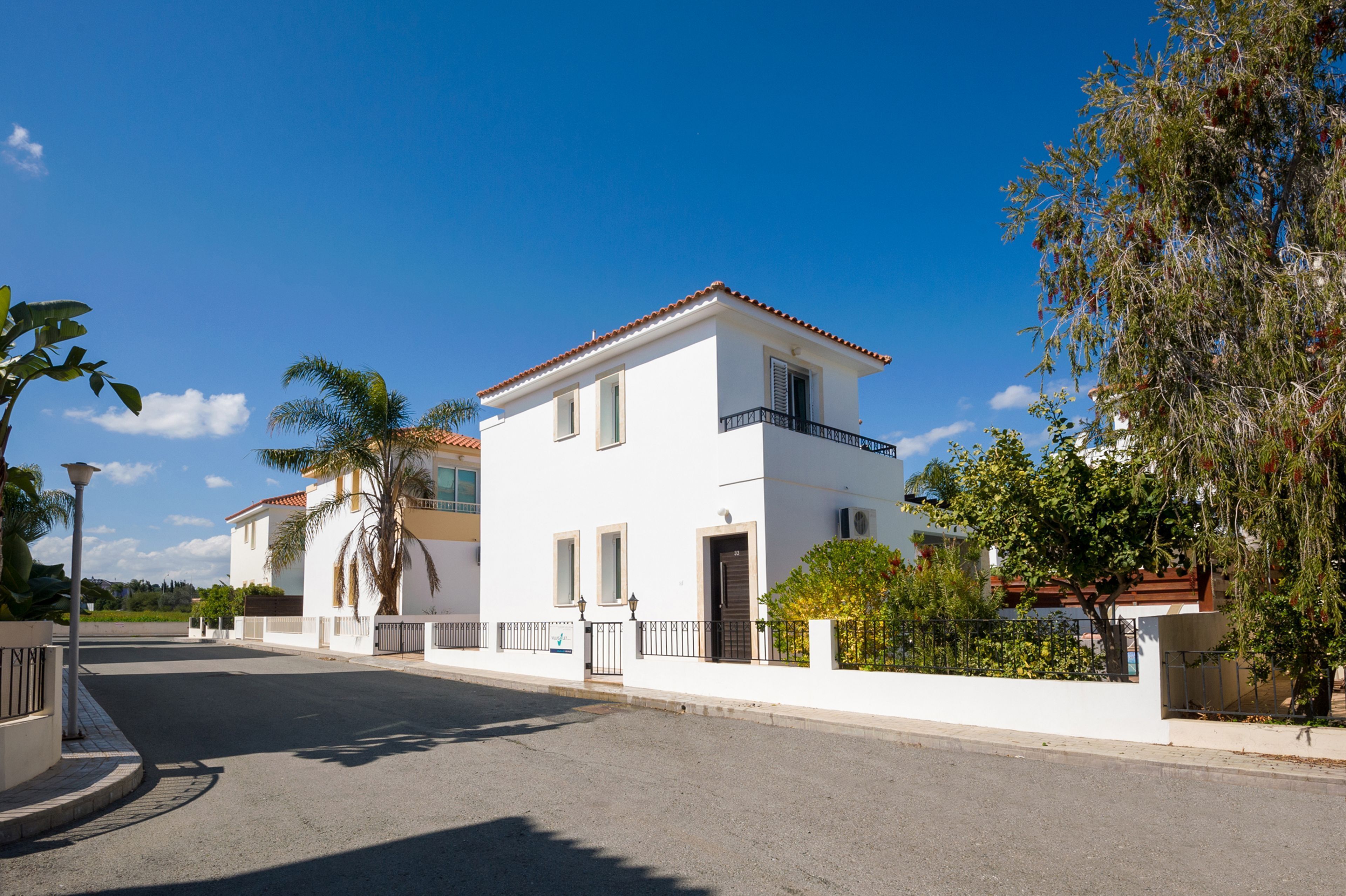 Casa blanca de dos pisos con balcón, rodeada de follaje y un cielo azul despejado.