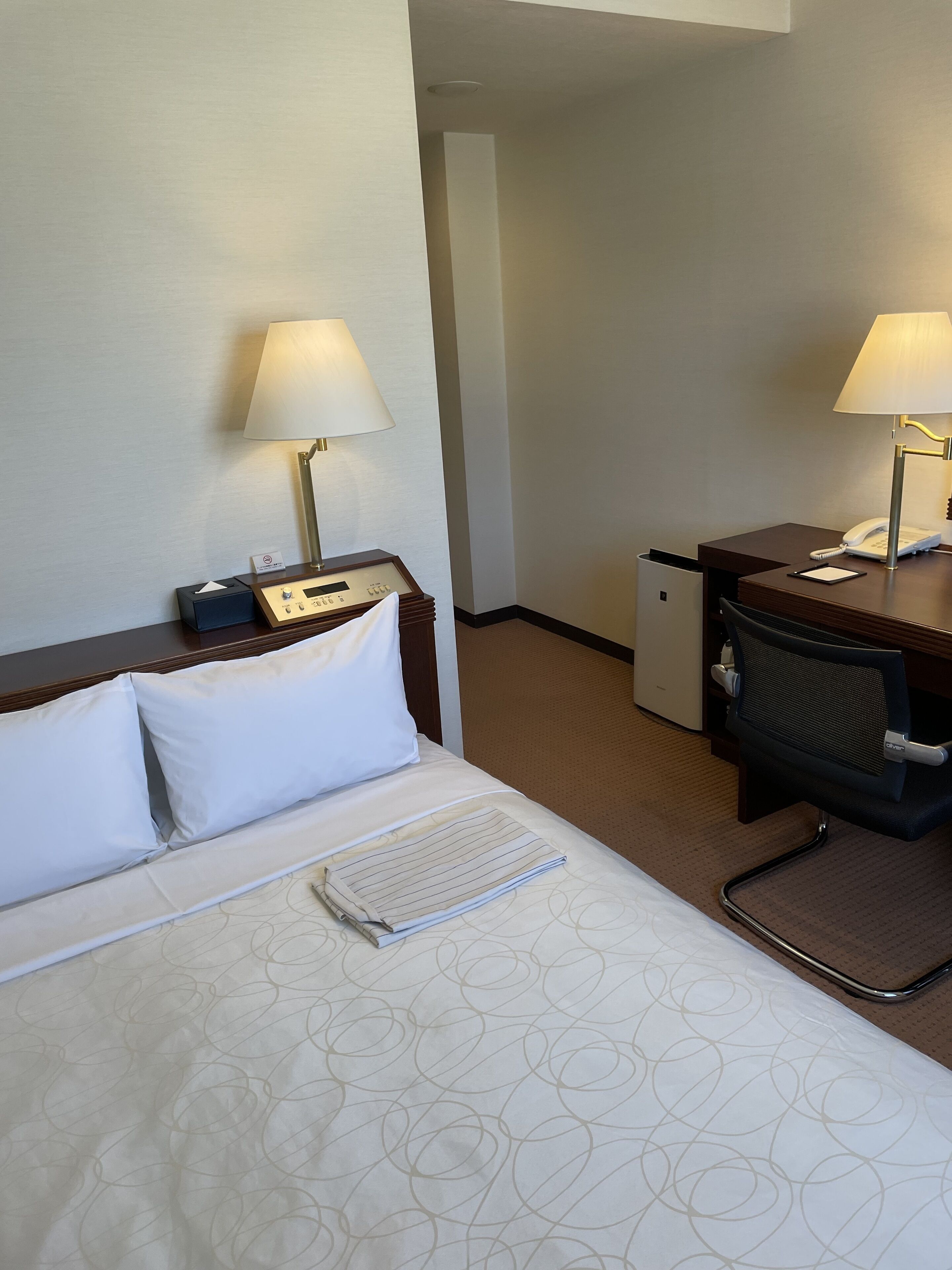 A hotel room with a bed, two bedside lamps, a desk with a telephone, and a chair.