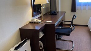 Ein Hotelzimmer mit Schreibtisch, Stuhl, Fernseher, Lampe, Telefon, Mülleimer und einem Fenster mit Vorhängen.