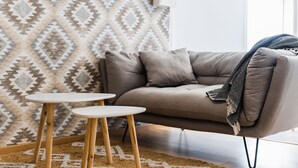 Un salon moderne avec un canapé beige, deux petites tables en bois et un mur orné d’un motif.