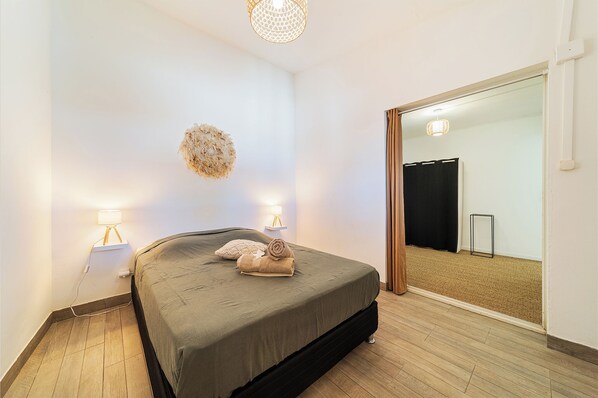 A bedroom with a bed, bedside lamps, a wall-mounted decorative piece, and a large mirror reflecting a door and a curtain.