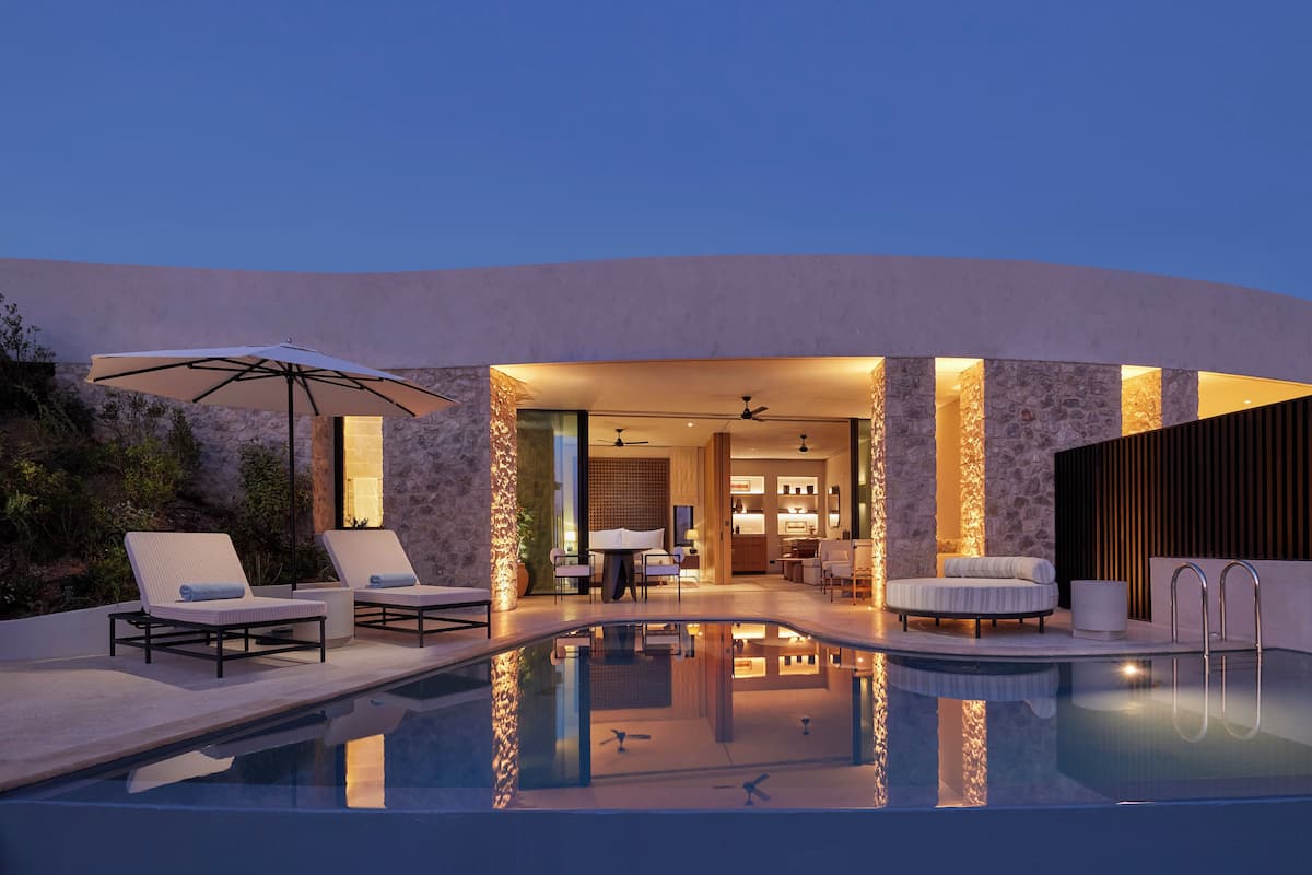 Zona de piscina exterior moderna amb gandules, una taula i un espai interior ben il·luminat visible a través d'una porta oberta.
