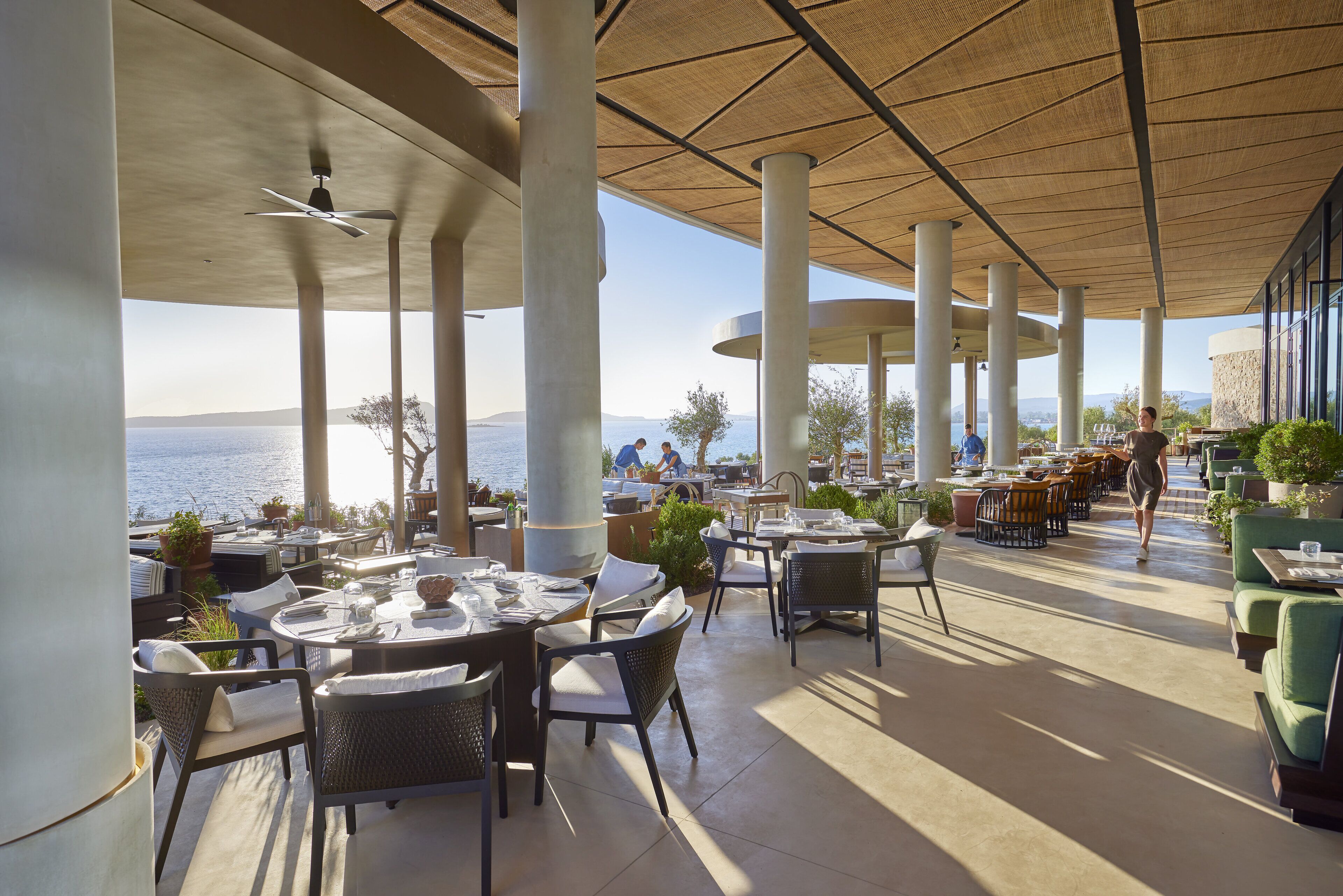 Un espacio de comedor al aire libre con mesas y sillas, pilares grandes y vista al mar.