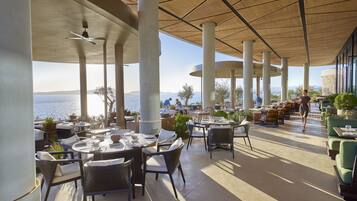 Un espacio de comedor al aire libre con mesas y sillas, pilares grandes y vista al mar.
