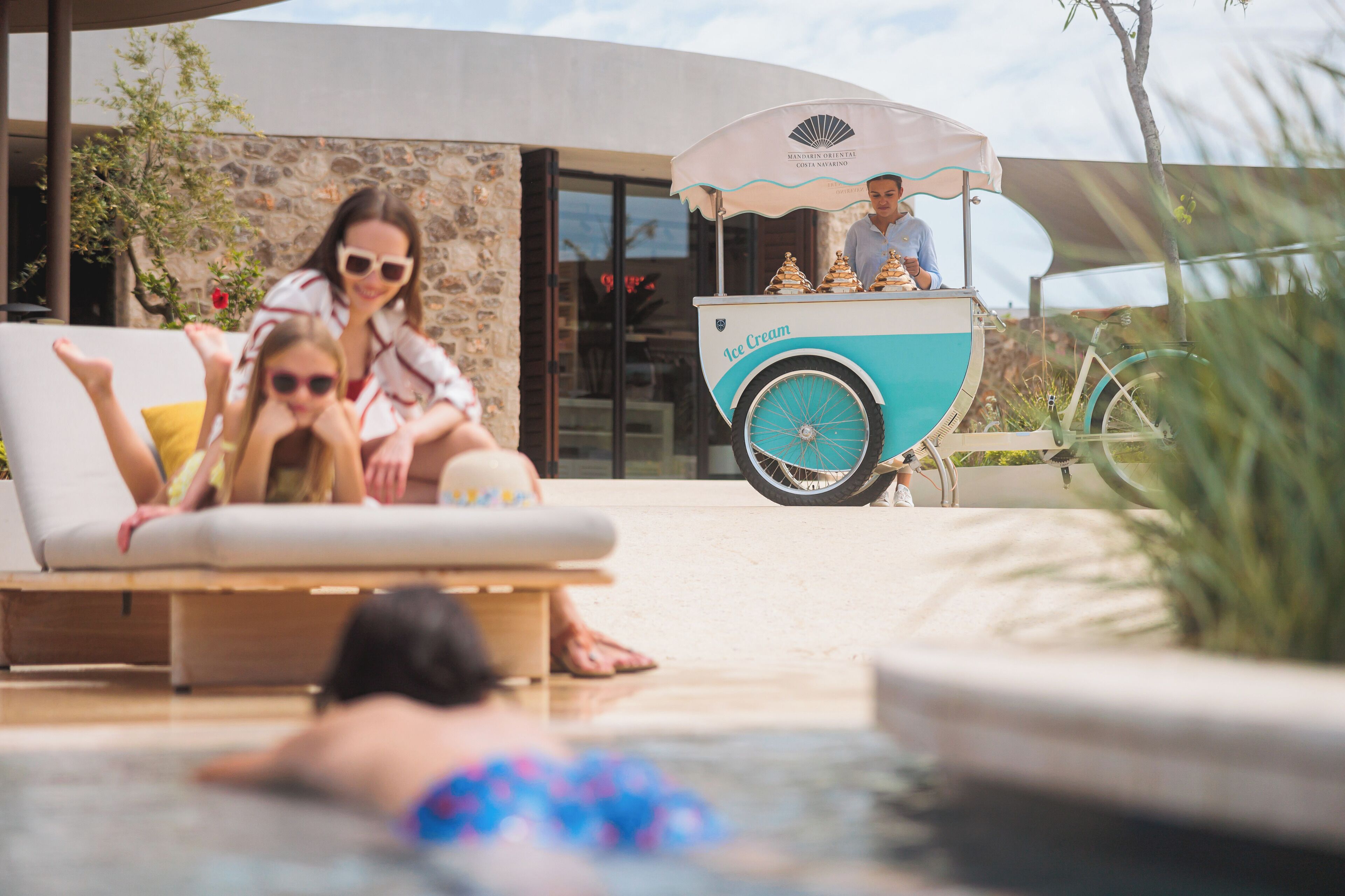 Un área junto a la piscina con sillones de descanso, una fuente de agua y un carrito de helados.
