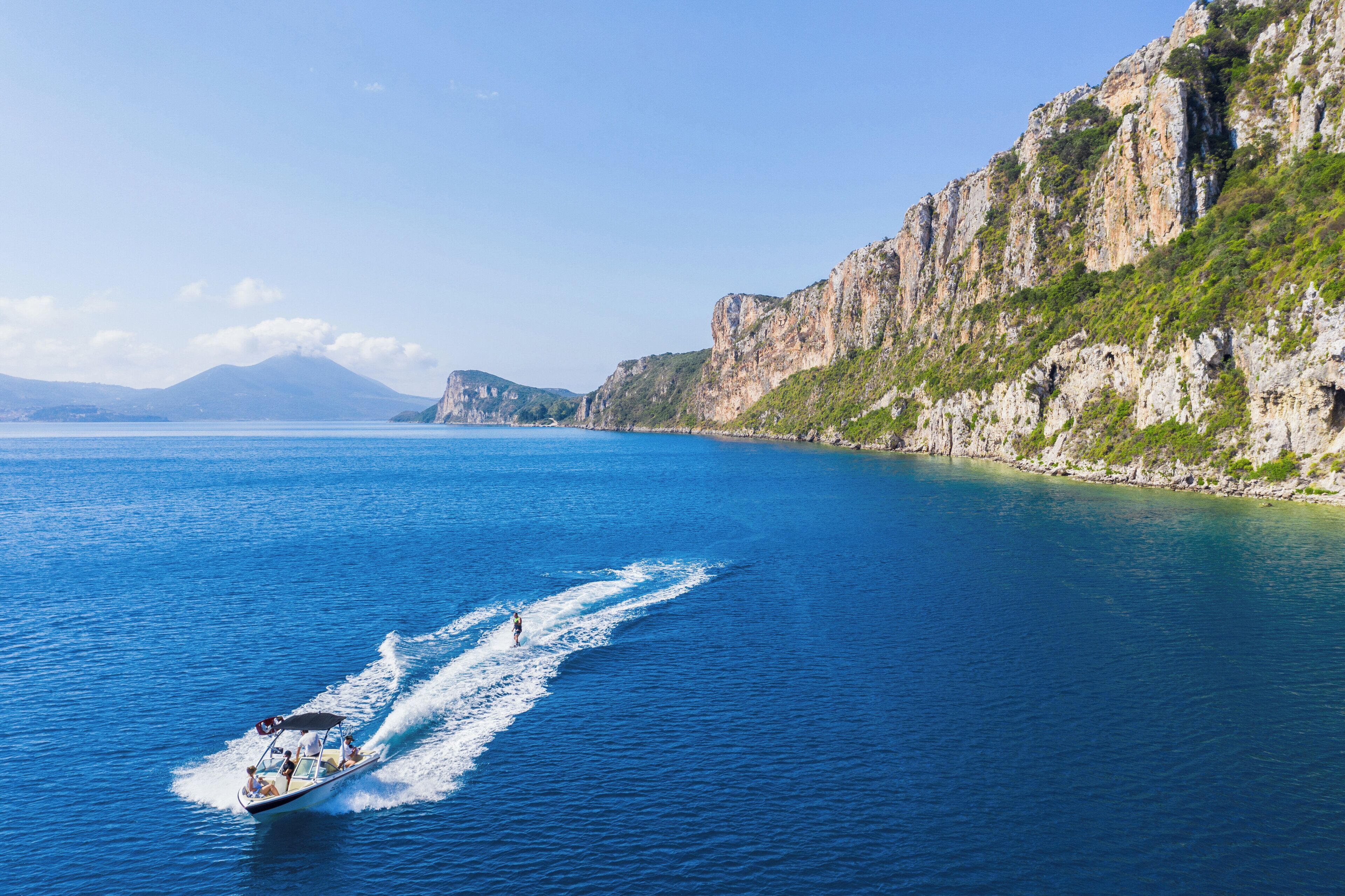 A boat on a calm sea with a rocky coastline and mountains in the background.