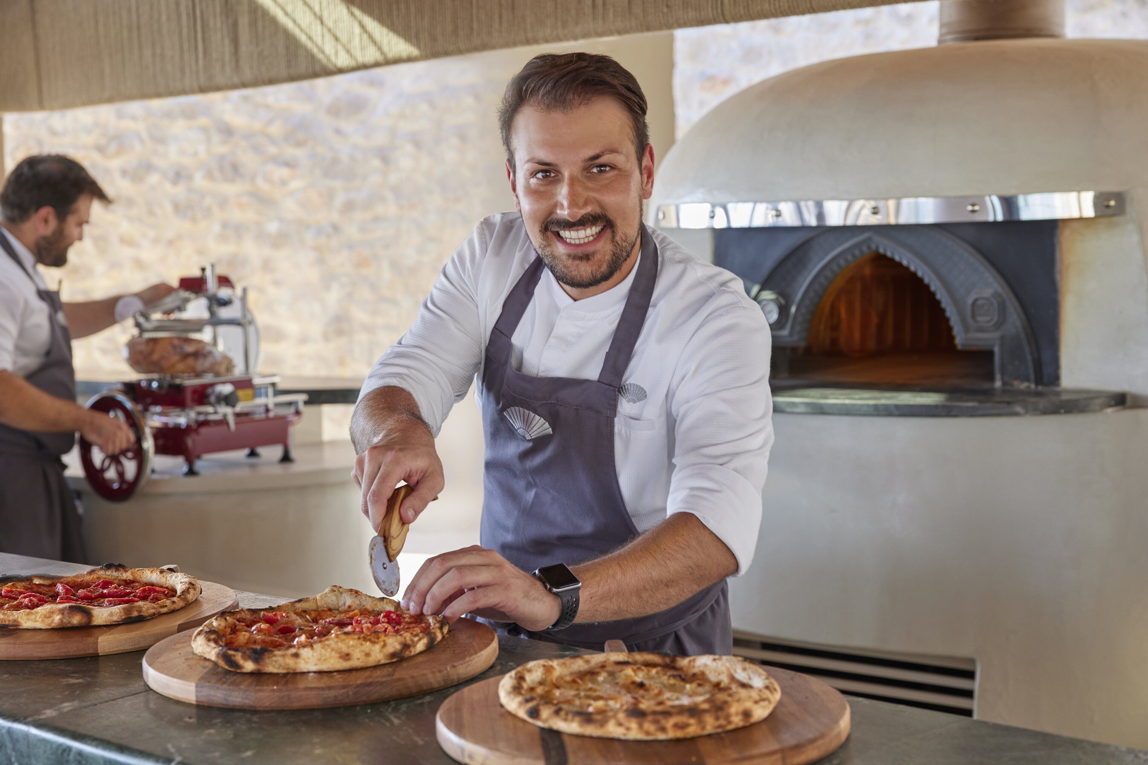 A chef preparing pizzas in a kitchen with a wood-fired oven.