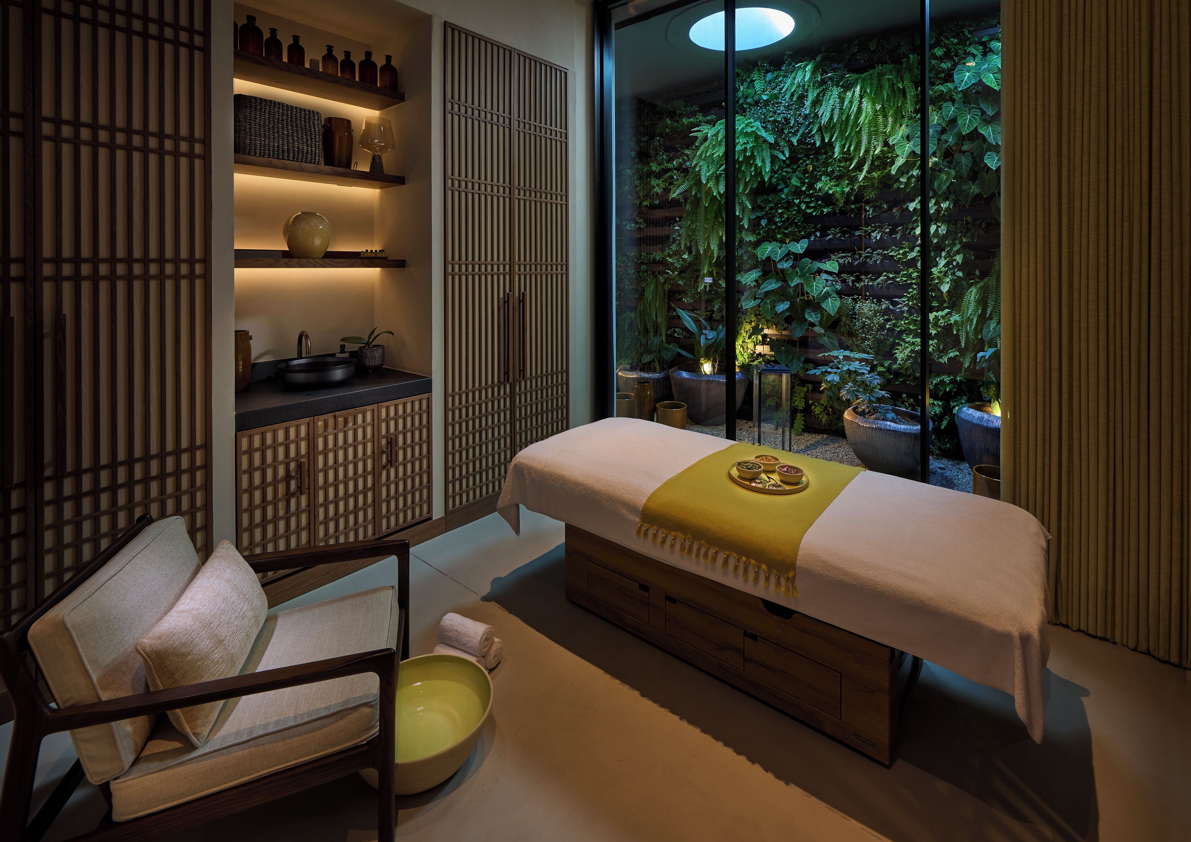 Une salle de spa équipée d’une table de massage, d’un fauteuil, d’une étagère avec des bouteilles et offrant une vue sur la verdure grâce à de grandes fenêtres.