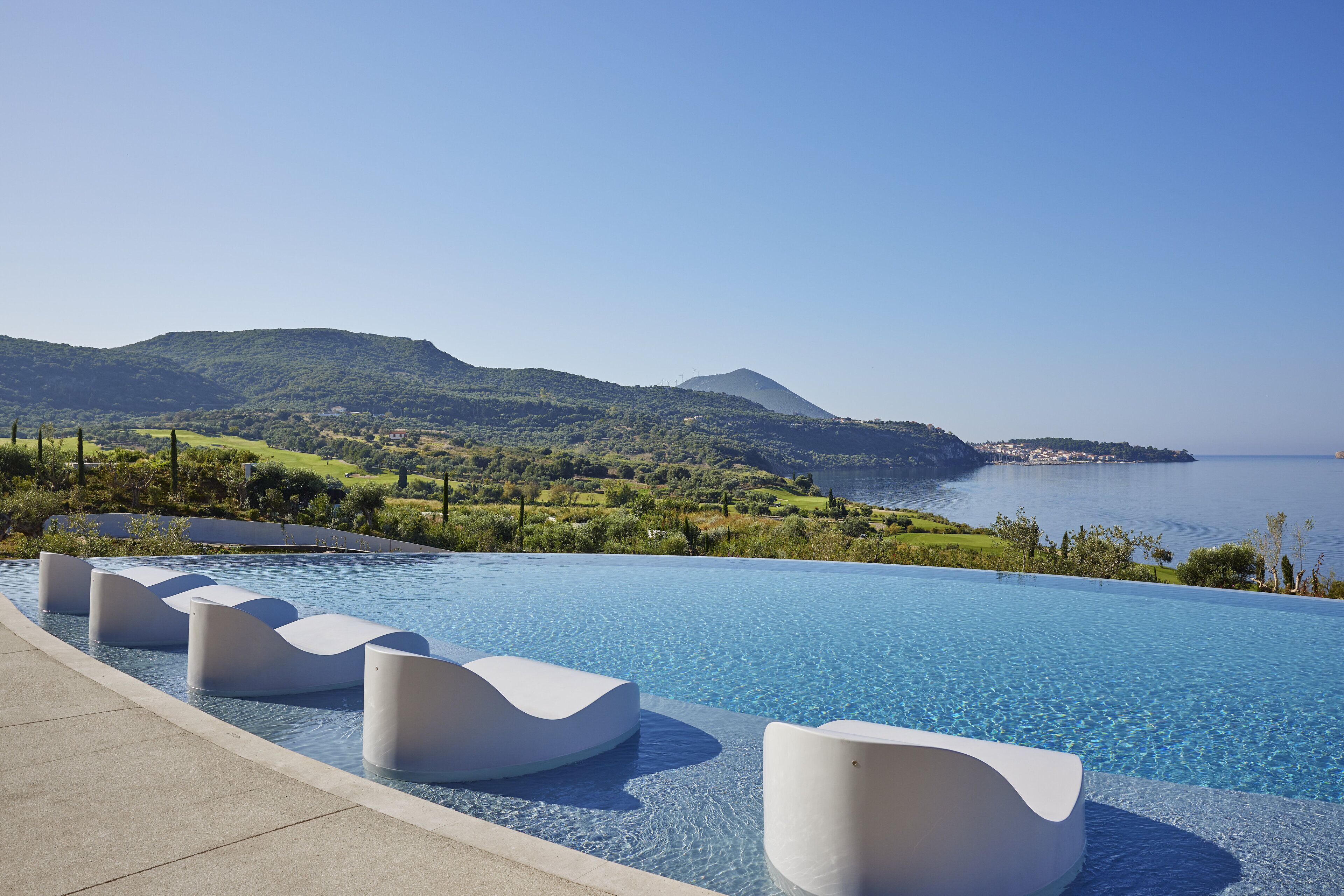 An infinity pool with modern white seating, surrounded by a scenic view of hills and the sea.
