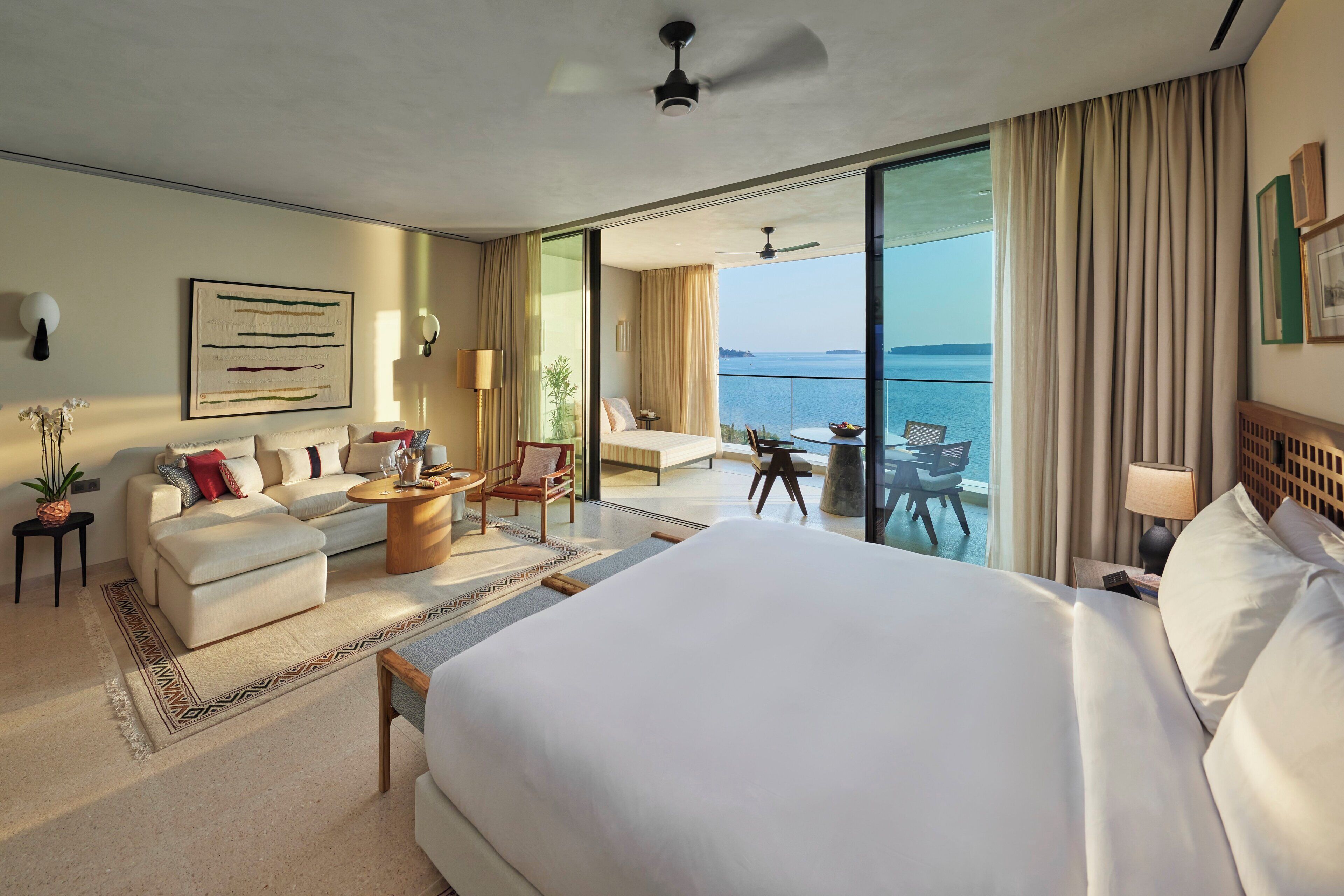 Habitación de hotel con una cama grande, un sofá, una mesa de comedor y un balcón con vista al mar.