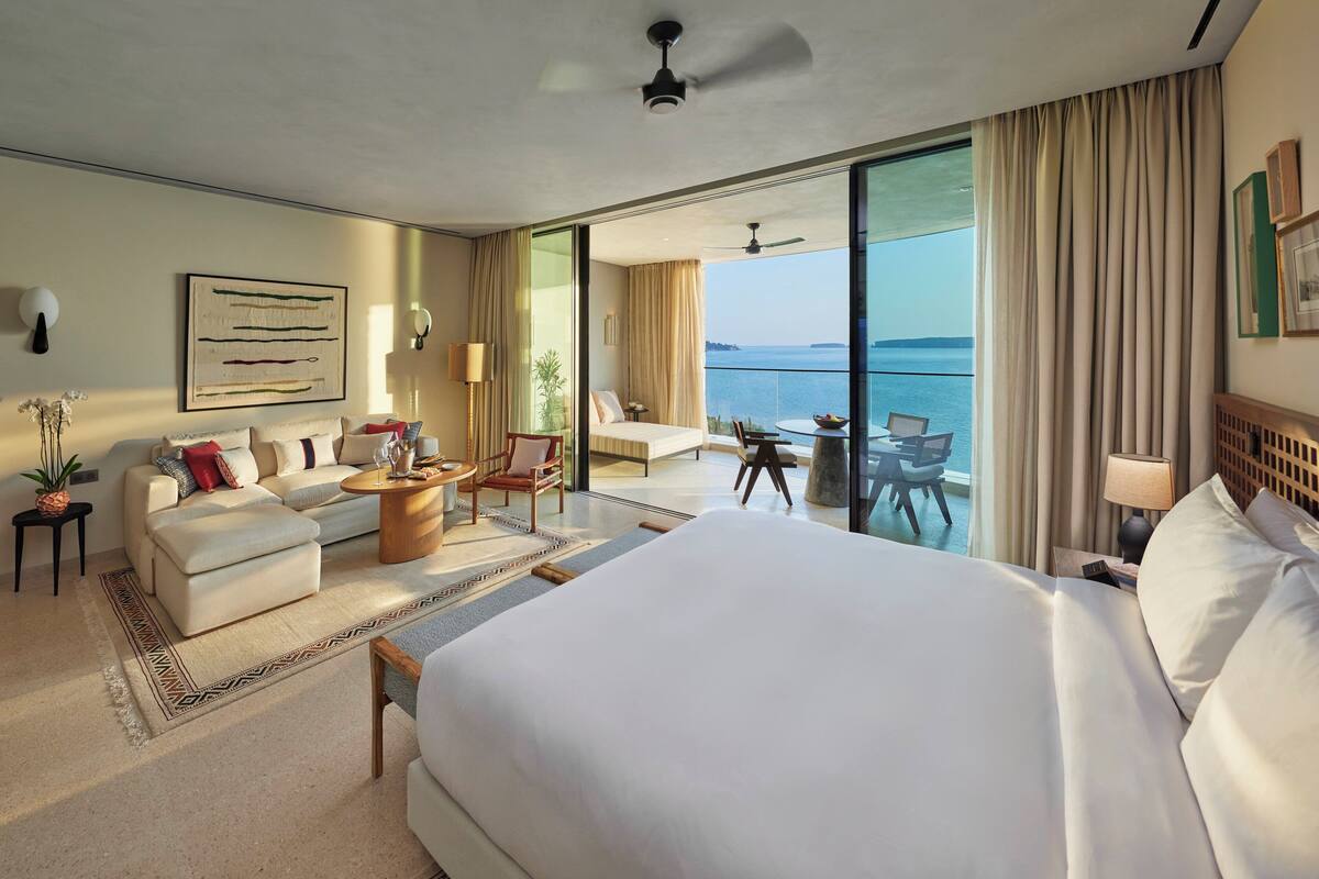 Amplia habitación de hotel con una cama grande, un sofá, una mesa de comedor y un balcón con vista al mar.