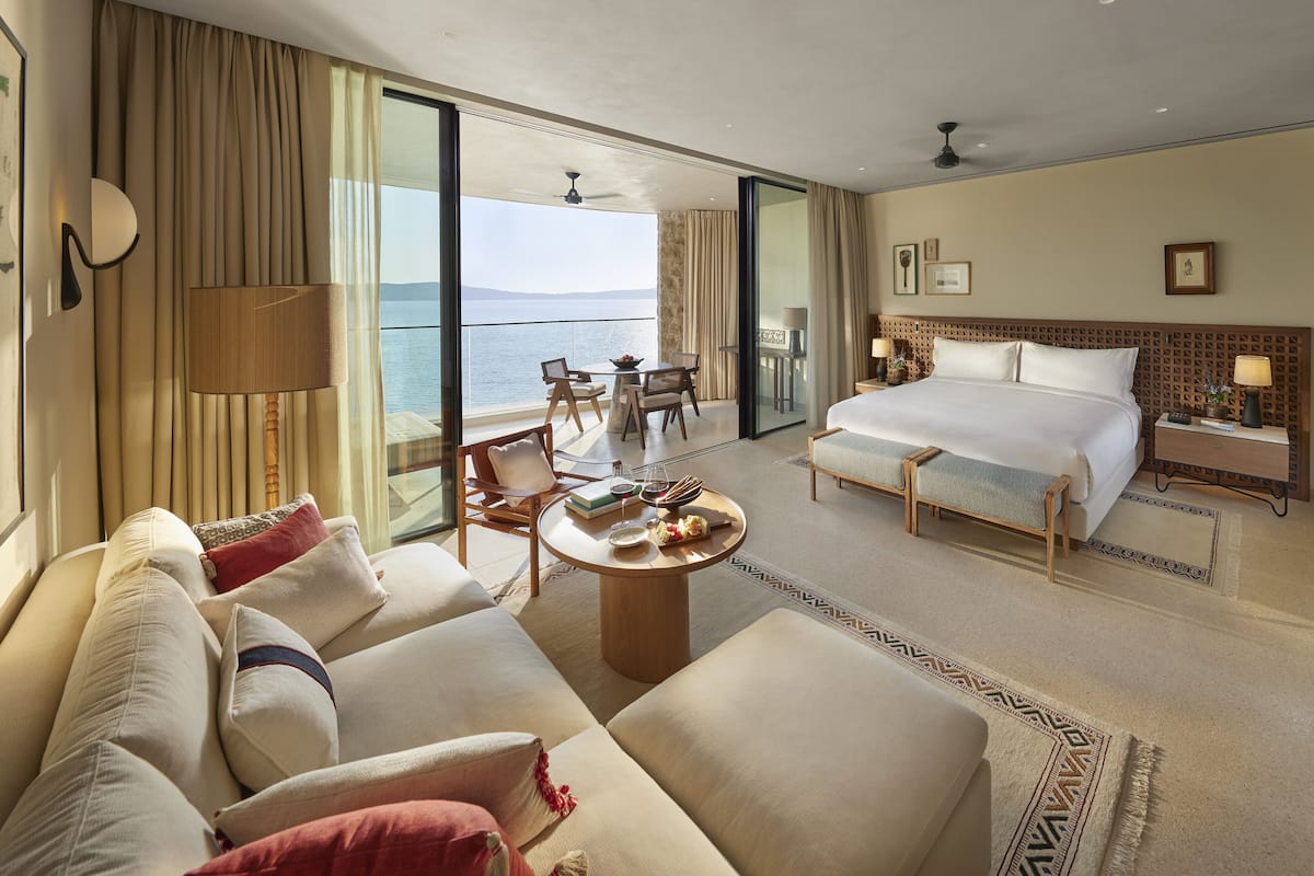 Habitación de hotel con una cama grande, un sofá, una mesa de comedor y un balcón con vistas al mar.