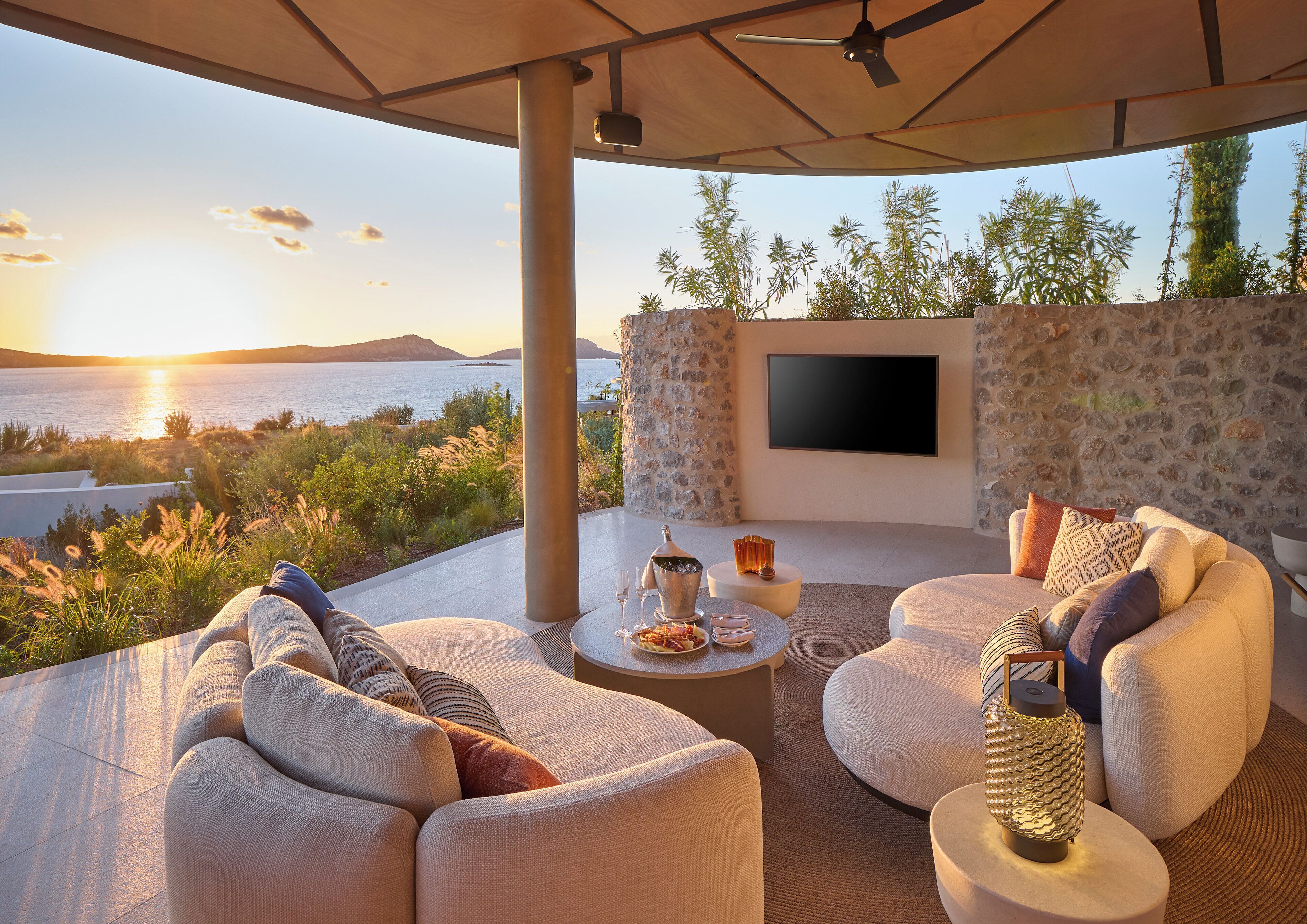 A modern outdoor seating area with a view of the sea at sunset, featuring comfortable seating and a flat-screen TV.