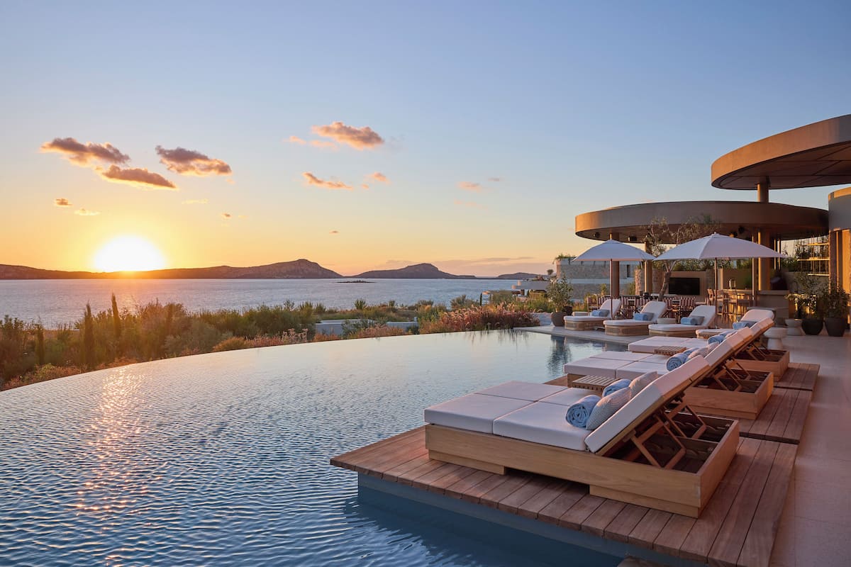 Una piscina infinita con sillas de descanso y sombrillas, con vistas a un cuerpo de agua y montañas distantes al atardecer.