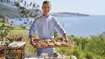 A person holding a tray with various dishes, standing in a dining area with a table set for a meal, overlooking a scenic coastal landscape.