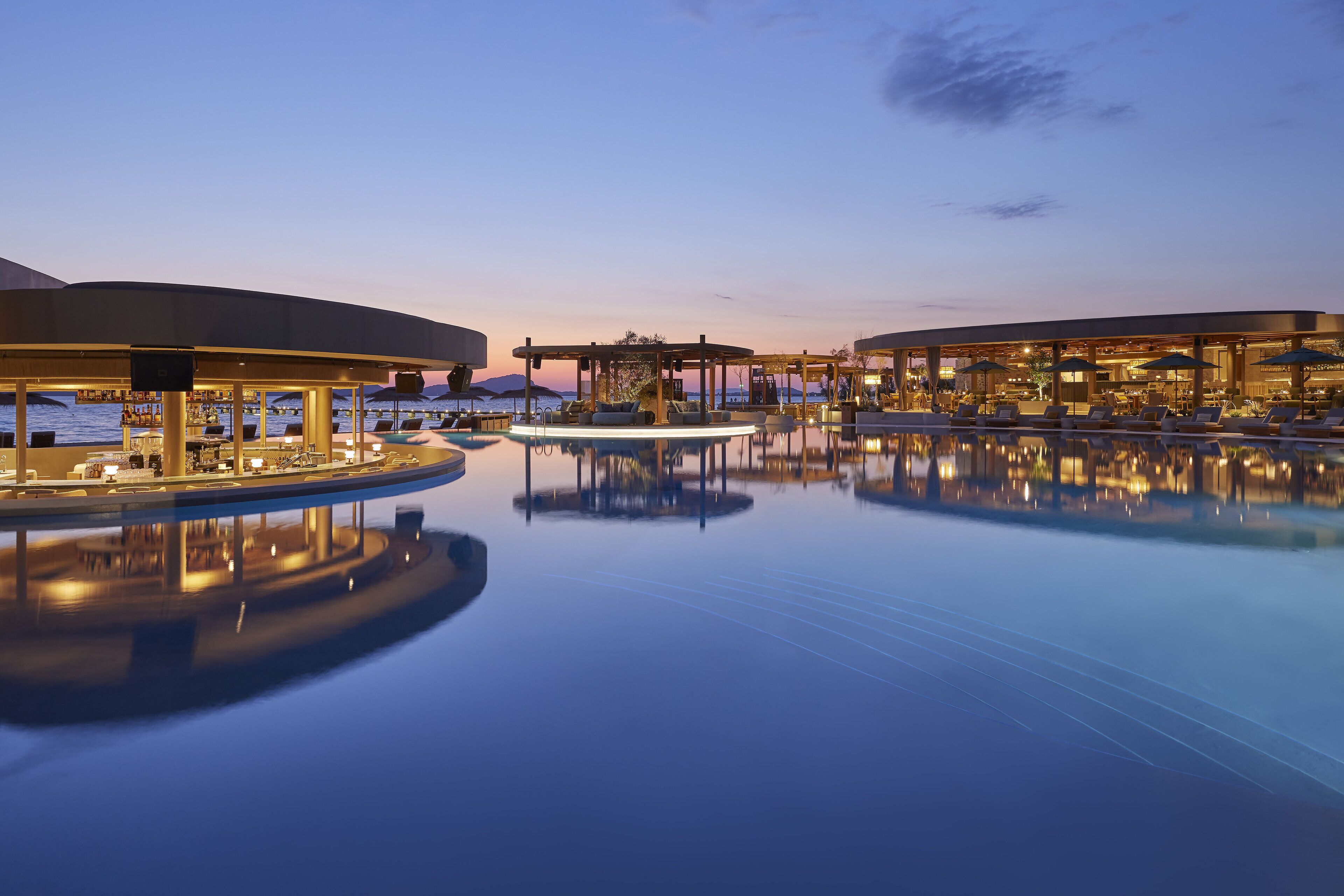 A serene pool area with lounge chairs and umbrellas, surrounded by well-lit buildings and a clear sky.