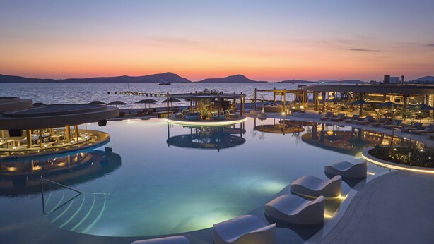 Zona de piscina en un resort con sillones de descanso y sombrillas, rodeada de edificios con terrazas y vistas al mar al atardecer.