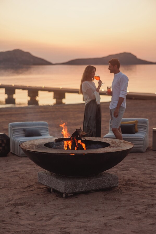 Una pareja junto a un fuego en un brasero en la playa, con sillones de descanso y vista al agua.