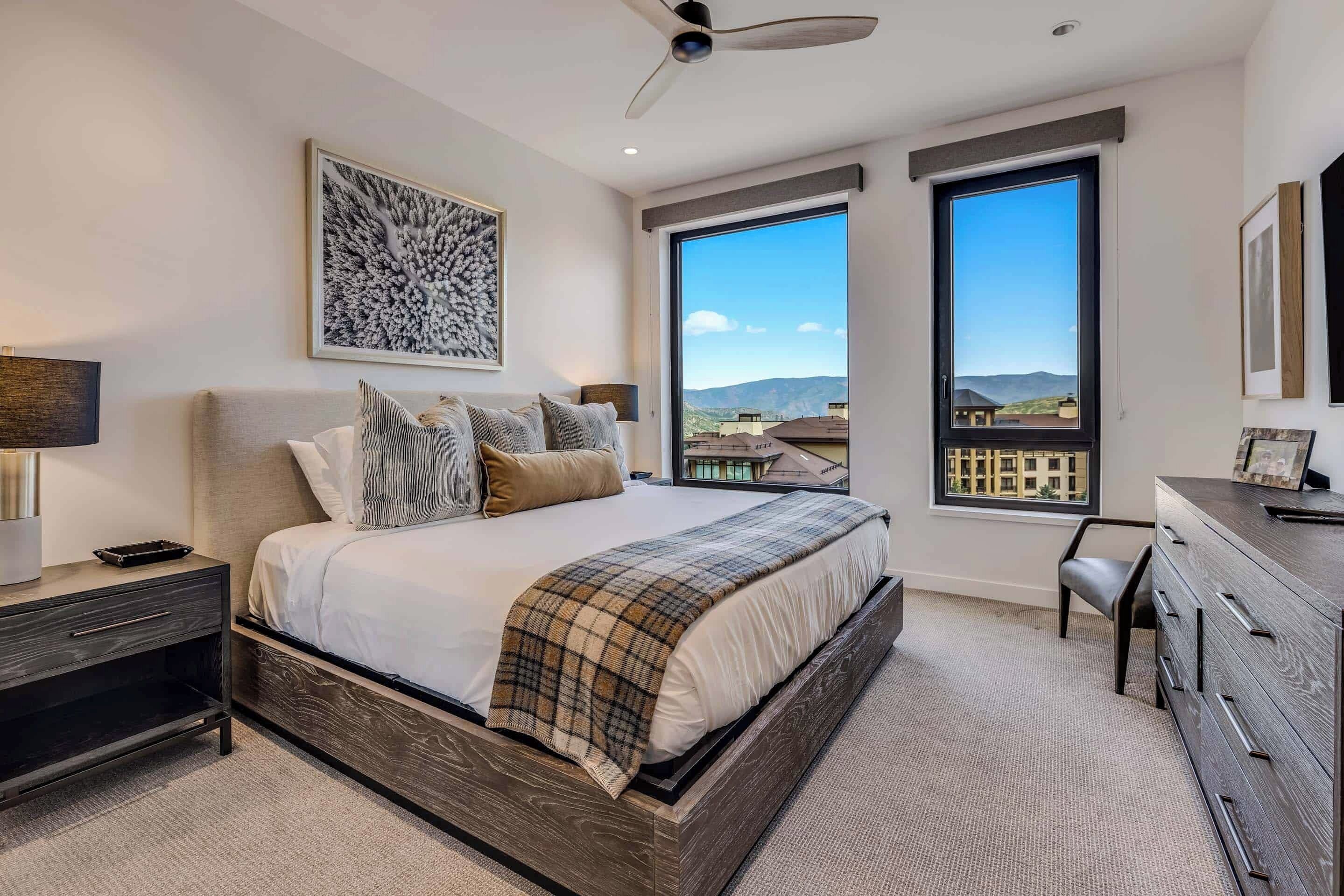 A modern bedroom with a large bed, a desk, and two windows offering a view of the cityscape.