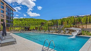 A swimming pool with lounge chairs and a waterfall feature, surrounded by a building and a mountainous landscape. - Electric Pass Lodge 508 (Snowmass Village)