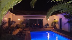 Une vue nocturne d’une maison avec une piscine, entourée de palmiers et de mobilier d’extérieur.
