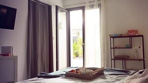 Une chambre d’hôtel avec un lit, une table de chevet avec une lampe, une étagère avec quelques objets, et une vue sur l’extérieur grâce à une porte coulissante.