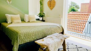 A bedroom with a bed, pillows, a bench, a dreamcatcher, a heart-shaped decoration, and a view of a red-tiled roof through a balcony door. - Charming lodging near the beach in De Koog (De Koog)
