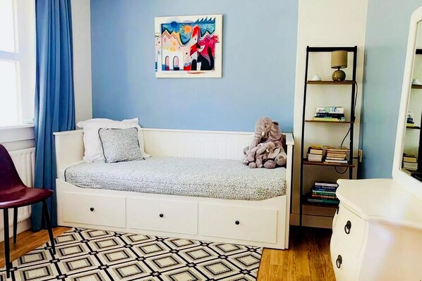 Un dormitorio con cama blanca, pared azul, un cuadro, estantería y alfombra estampada.