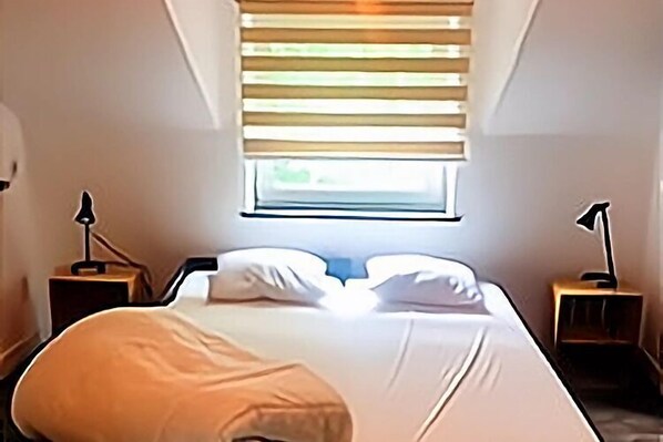 A neatly made bed with white linens, a window with blinds, and two bedside tables with lamps.