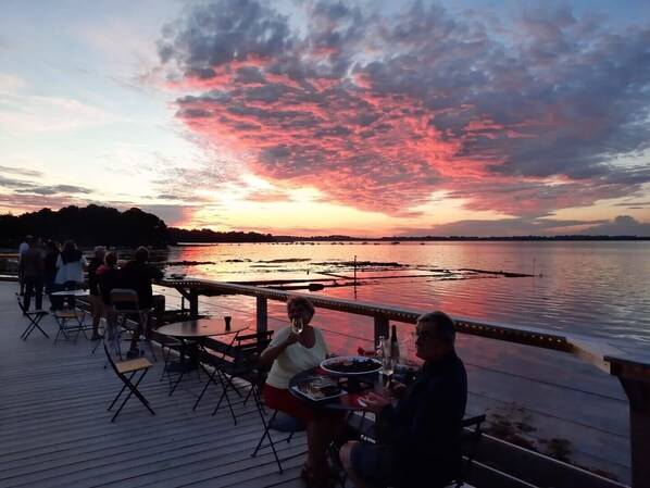 Une vue sur le coucher de soleil depuis une terrasse en bois donnant sur une étendue d’eau où se trouvent des bateaux, des tables et des chaises.