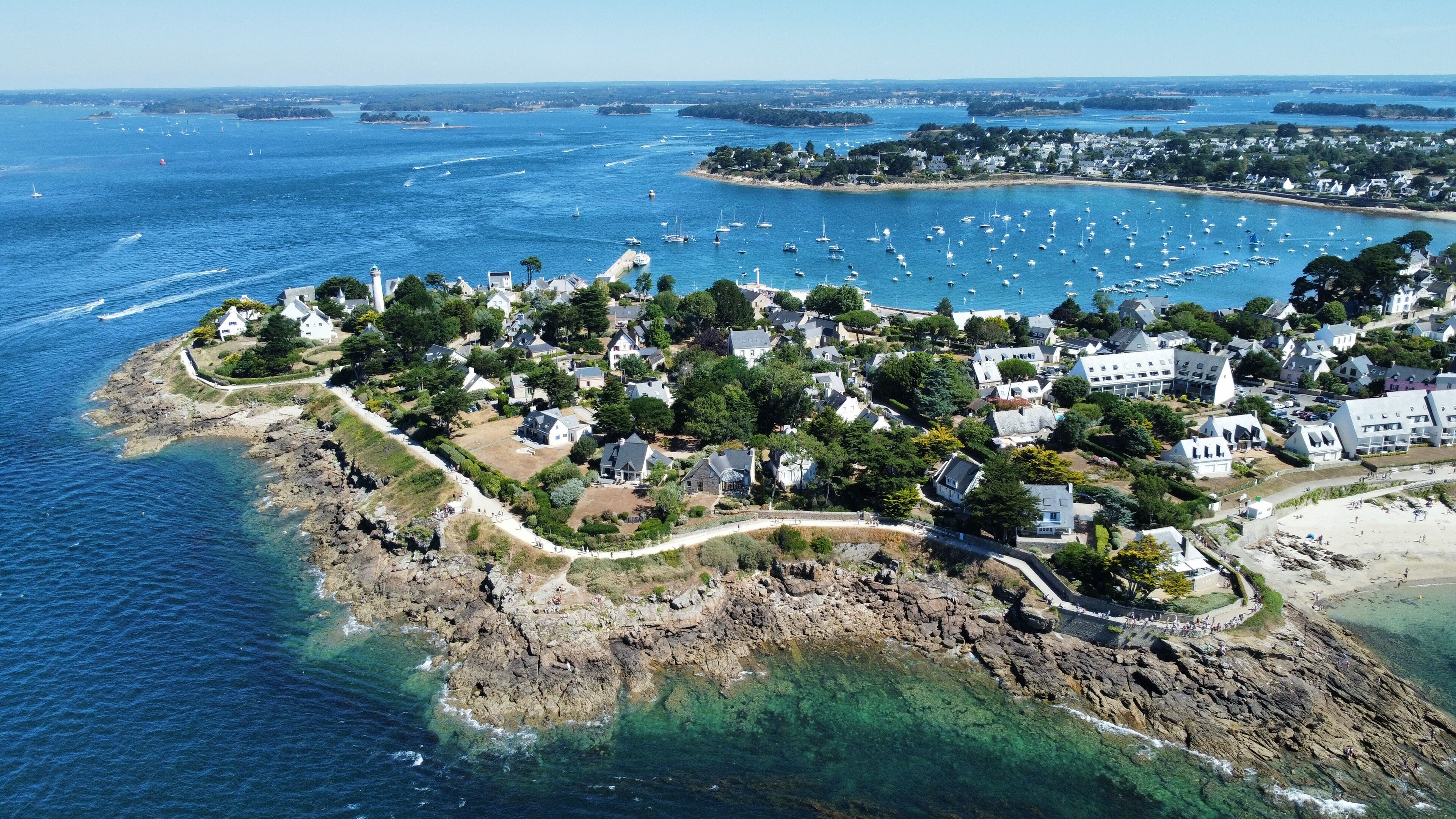 Ett flygbildsbild av en kuststad med hus längs strandlinjen, klart blått vatten och båtar i hamnen.