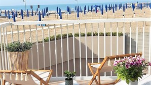 A balcony with a view of the beach, featuring a railing, two wooden chairs, a small table, and potted plants. - Beachside Apartment for 4 in Caorle (Porto Santa Margherita (VE))