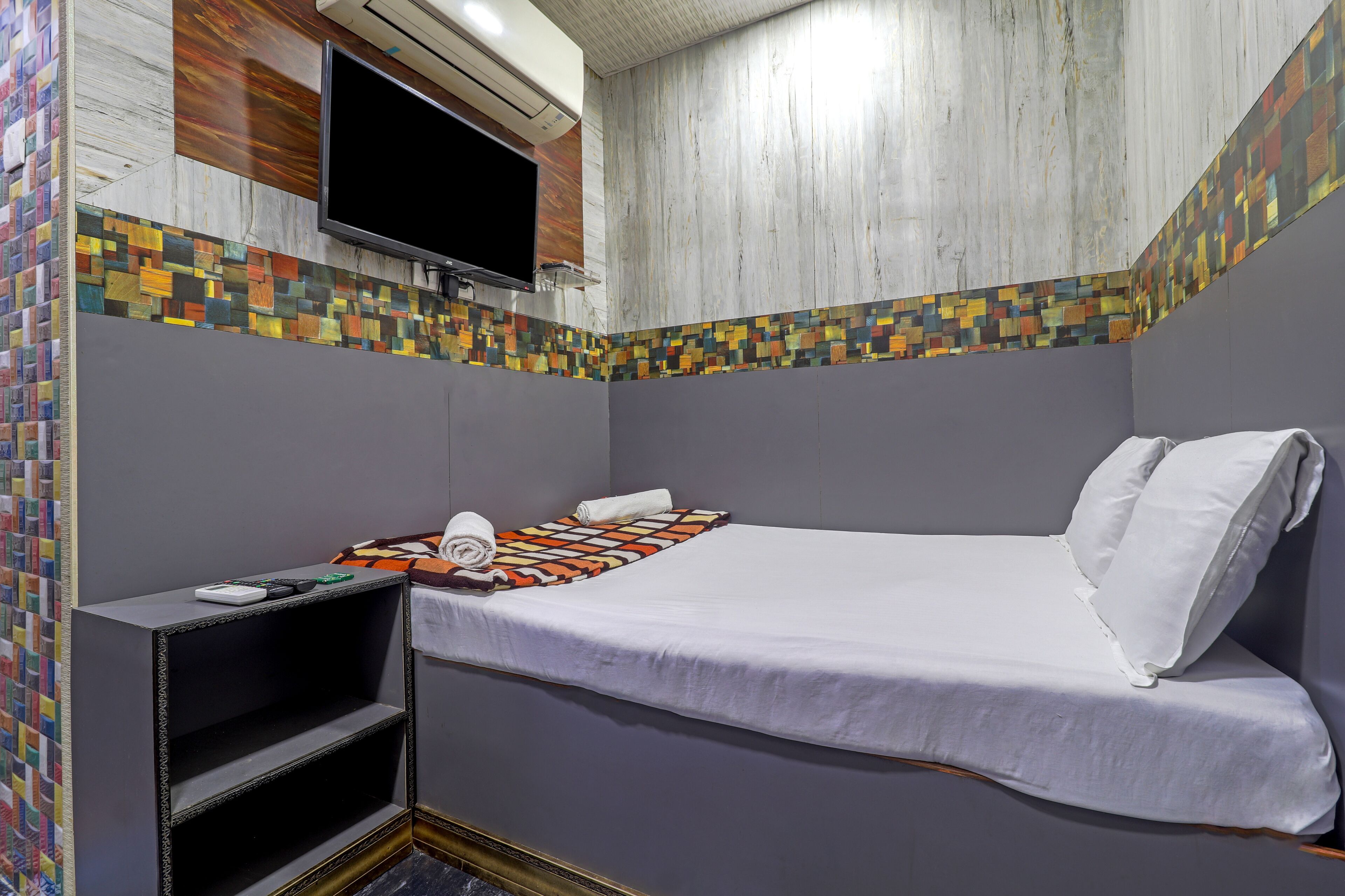 Un dormitorio pequeño y ordenado con cama, televisor montado en la pared y un llamativo borde de mosaicos.